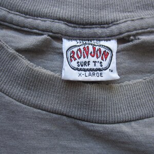 Op de afbeelding: Close-up van een grijs t-shirt met een wit label. Het label leest "RON JON SURF T'S" in rode en zwarte letters, met "X-LARGE" eronder gedrukt. Het shirt is gemaakt van een zachte, getextureerde stof.