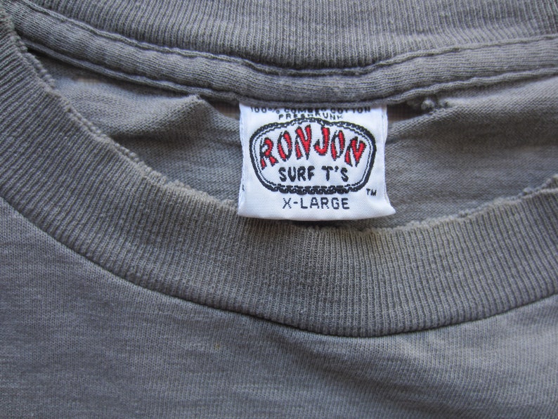 Op de afbeelding: Close-up van een grijs t-shirt met een wit label. Het label leest "RON JON SURF T'S" in rode en zwarte letters, met "X-LARGE" eronder gedrukt. Het shirt is gemaakt van een zachte, getextureerde stof.