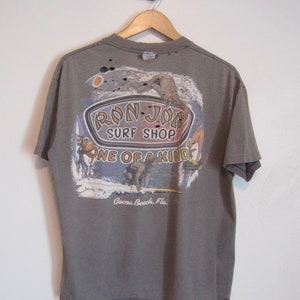 Op de afbeelding: Een grijs t-shirt met een afbeelding van Ron Jon Surf Shop. De afbeelding toont surfers, golven en de tekst "Ron Jon Surf Shop One of a Kind Cocoa Beach, Fla.". Het shirt hangt aan een houten hanger.