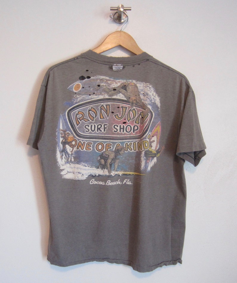 Op de afbeelding: Een grijs t-shirt met een afbeelding van Ron Jon Surf Shop. De afbeelding toont surfers, golven en de tekst "Ron Jon Surf Shop One of a Kind Cocoa Beach, Fla.". Het shirt hangt aan een houten hanger.