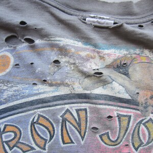 Op de afbeelding: Verweerd grijs t-shirt met een surferafbeelding en de tekst "RON JON". Het shirt heeft meerdere gaten en een vintage uitstraling. De afbeelding bevat oranje, blauwe en roze accenten.