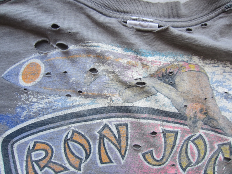 Op de afbeelding: Verweerd grijs t-shirt met een surferafbeelding en de tekst "RON JON". Het shirt heeft meerdere gaten en een vintage uitstraling. De afbeelding bevat oranje, blauwe en roze accenten.