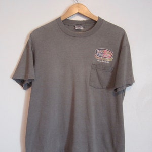 Op de afbeelding: Een grijs t-shirt met een zak en de tekst "RON JON SURF SHOP" in een kleurrijk logo. De tekst bevat ook "ONE OF A KIND" en "Cocoa Beach, FL.". Het t-shirt hangt aan een houten hanger tegen een witte muur.