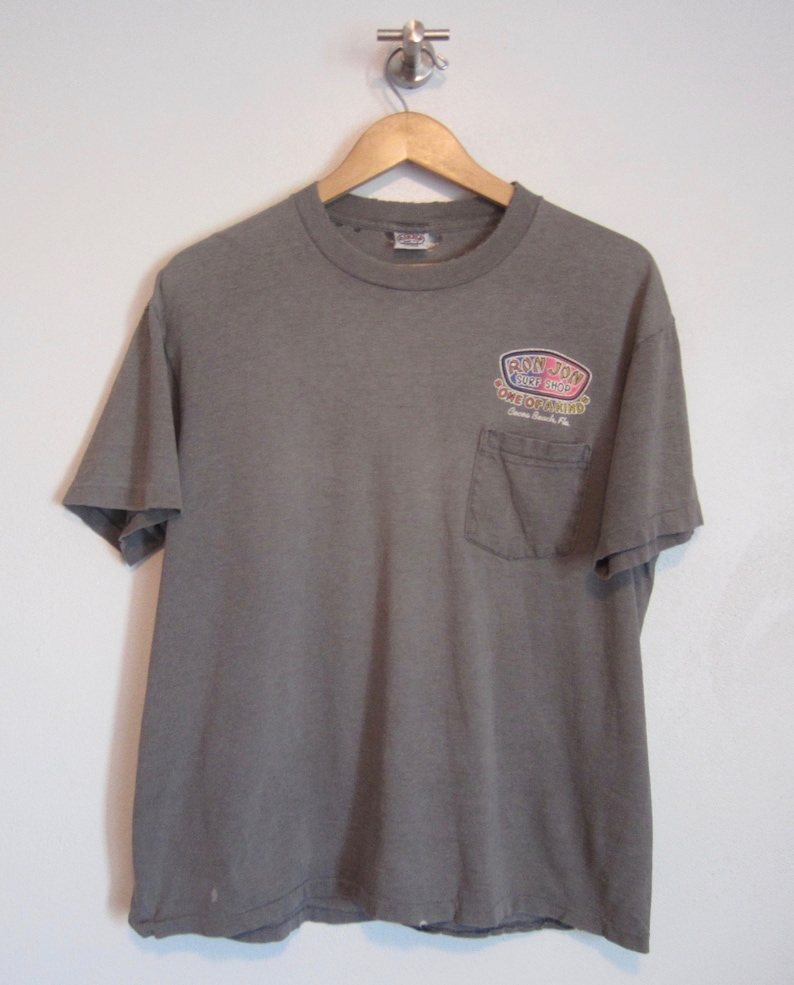 Op de afbeelding: Een grijs t-shirt met een zak en de tekst "RON JON SURF SHOP" in een kleurrijk logo. De tekst bevat ook "ONE OF A KIND" en "Cocoa Beach, FL.". Het t-shirt hangt aan een houten hanger tegen een witte muur.