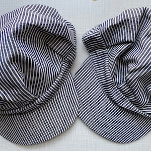 May include: Two navy and white striped hats, each with a rounded crown and a short peak. The hats are made of a woven fabric with vertical stripes. The style is similar to a railway worker's cap.