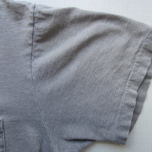 Op de afbeelding: Close-up van een grijze t-shirtmouw met een zak. De shirt heeft een afbeelding met de woorden "Spoon Shop" en "Kind" in een roze en geel ontwerp. De tekst "Fla." is ook zichtbaar.