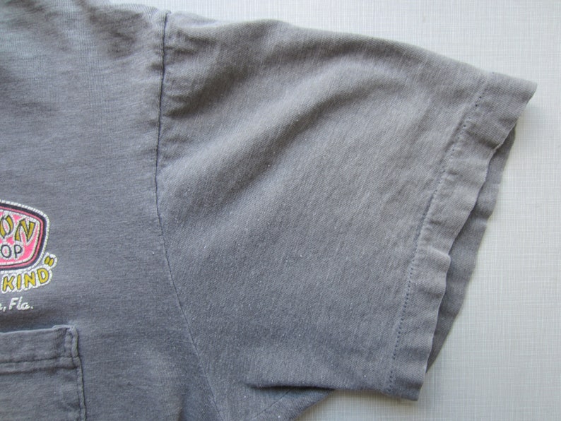 Op de afbeelding: Close-up van een grijze t-shirtmouw met een zak. De shirt heeft een afbeelding met de woorden "Spoon Shop" en "Kind" in een roze en geel ontwerp. De tekst "Fla." is ook zichtbaar.