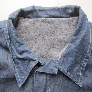 Puede incluir: Primer plano de una chaqueta vaquera azul con forro interior gris y difuso. El cuello está doblado hacia abajo y la chaqueta tiene costuras blancas a lo largo de las costuras. La chaqueta está abotonada en la parte superior.