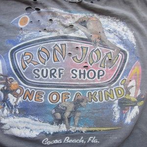 Op de afbeelding: Grijze t-shirt met een verweerde afbeelding van surfers, surfplanken en de tekst "RON JON SURF SHOP ONE OF A KIND Cocoa Beach, Fla." Het ontwerp bevat blauwe, oranje en gele accenten, met een vintage, versleten uitstraling.