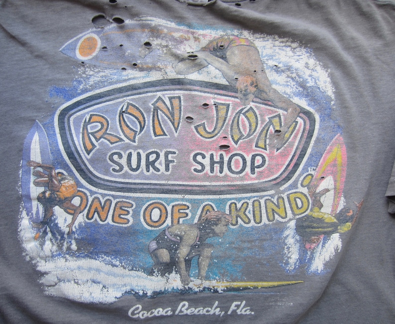 Op de afbeelding: Grijze t-shirt met een verweerde afbeelding van surfers, surfplanken en de tekst "RON JON SURF SHOP ONE OF A KIND Cocoa Beach, Fla." Het ontwerp bevat blauwe, oranje en gele accenten, met een vintage, versleten uitstraling.