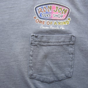 Op de afbeelding: Grijze t-shirt met een zak en een kleurrijk "Ron Jon Surf Shop" logo. Het logo bevat de tekst "One of a Kind" en "Cocoa Beach, Fla."