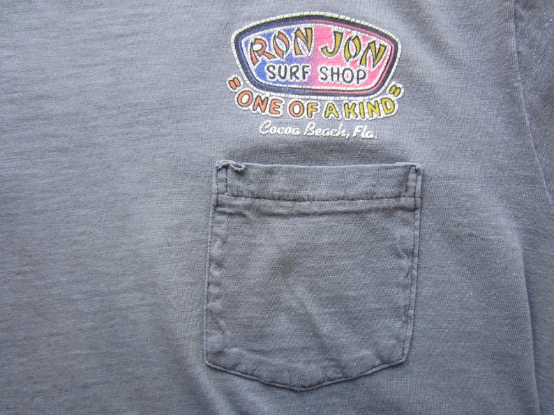 Op de afbeelding: Grijze t-shirt met een zak en een kleurrijk "Ron Jon Surf Shop" logo. Het logo bevat de tekst "One of a Kind" en "Cocoa Beach, Fla."