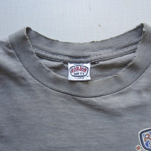 Op de afbeelding: Close-up van een vervaagd grijs t-shirt met een versleten halslijn. De shirt heeft een "RON JON SURF T'S" label en een kleurrijke afbeelding aan de onderkant. Het label geeft een maat X-Large aan.