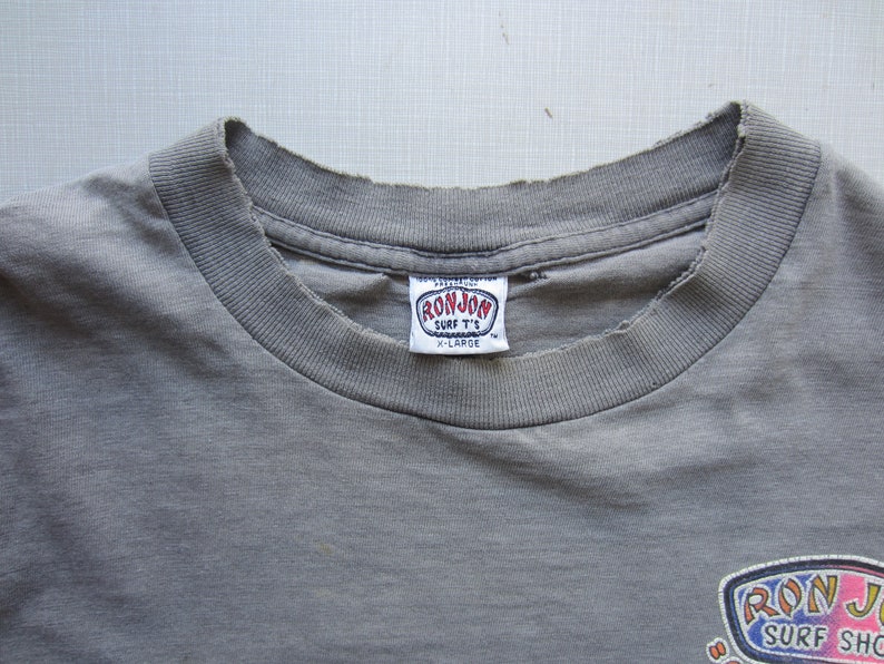 Op de afbeelding: Close-up van een vervaagd grijs t-shirt met een versleten halslijn. De shirt heeft een "RON JON SURF T'S" label en een kleurrijke afbeelding aan de onderkant. Het label geeft een maat X-Large aan.