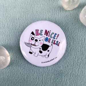 May include: A white button featuring a black and white cartoon dog holding a knife. The text "Be Nice! Or Else" is written in rainbow colours.