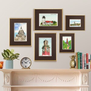 May include: A collection of five framed watercolor illustrations of various buildings and landmarks. The frames are brown with gold trim and are arranged on a white wall above a white mantelpiece. The mantelpiece has a clock, a potted plant, and a pineapple on it.