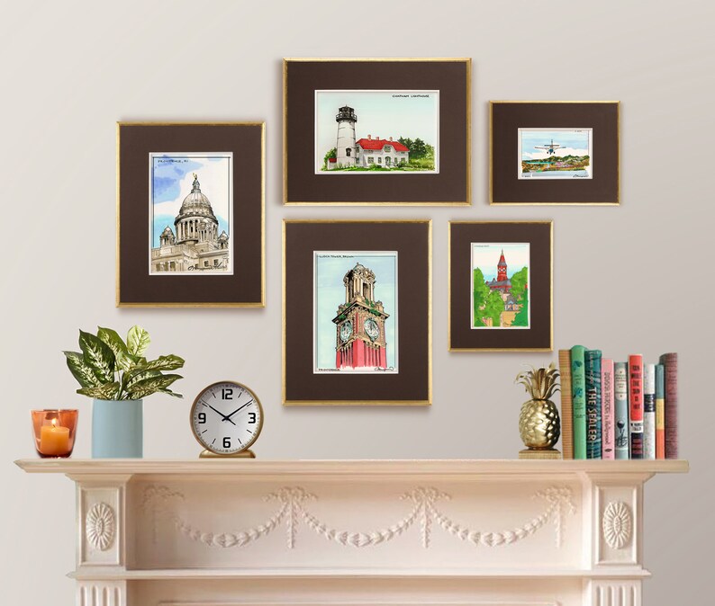 May include: A collection of five framed watercolor illustrations of various buildings and landmarks. The frames are brown with gold trim and are arranged on a white wall above a white mantelpiece. The mantelpiece has a clock, a potted plant, and a pineapple on it.