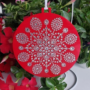 May include: A red, circular Christmas bauble featuring a rhinestone snowflake design. It has a white ribbon for hanging and is set against a background of green foliage and red flowers. The bauble is approximately 10 cm in diameter.
