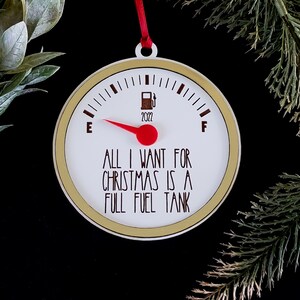 May include: A white Christmas ornament shaped like a gas gauge with a red needle pointing to full. The gauge reads "All I want for Christmas is a full fuel tank".