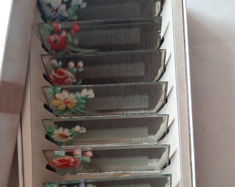 May include: A set of ten glass place card holders with painted floral designs. Each holder features a different floral arrangement with red, yellow, blue, and green flowers.