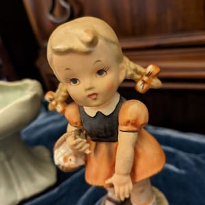 Op de afbeelding: Een porseleinen beeldje van een meisje met blonde vlechten, gekleed in een zwart en oranje jurkje. Ze houdt een mandje vast met een vogeltje erin. Het beeldje staat op een voet met een bloemenmotief.