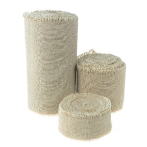 May include: Three rolls of natural beige burlap ribbon, each with a frayed edge. The rolls are stacked on top of each other, with the largest roll on the bottom and the smallest roll on top.