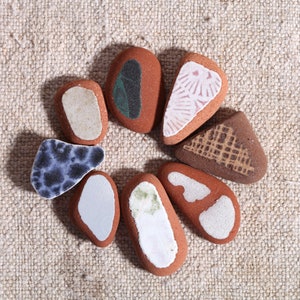 May include: A selection of terracotta-coloured, irregularly shaped stones, each with a unique design. Some stones feature white, blue, or green patterns, while others have textured brown designs. The stones are arranged in a circular pattern.