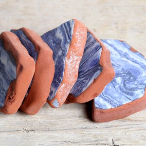 Puede incluir: Un juego de cuatro posavasos de cerámica planos e irregulares con un patrón jaspeado en azul y blanco en la parte superior, enmarcados por un borde grueso de arcilla color terracota sin esmaltar con textura rugosa y acabado mate.