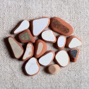 May include: A collection of fifteen smooth, rounded stones with flat surfaces. Each stone features a terracotta-colored base with distinct, contrasting inlays in shades of white, cream, tan, dark brown, and charcoal gray, arranged in a cluster.