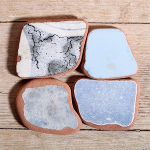 Puede incluir: Cuatro posavasos de cerámica de forma irregular con borde marrón rojizo. Los posavasos tienen diferentes diseños: uno con un patrón agrietado blanco y gris, uno azul claro liso y dos con patrones moteados azul claro. Dispuestos sobre una superficie de madera.
