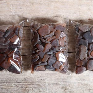 Op de afbeelding: Drie doorzichtige plastic zakken gevuld met zeeglas in tinten bruin, grijs en amber. De stukken zeeglas variëren in grootte en vorm. De zakken zijn gerangschikt op een lichtbruin houten oppervlak.