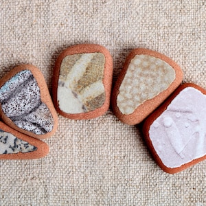 May include: A collection of six decorative stones with a reddish-brown border and a lighter-colored interior. The interior designs vary, featuring patterns of gray, beige, and white. The stones are arranged on a textured, beige surface.
