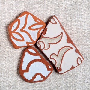 Puede incluir: Tres baldosas de cerámica color terracota con diseños decorativos blancos y marrones. Una baldosa es cuadrada, otra rectangular y la tercera triangular. Las baldosas están dispuestas sobre una superficie de lino beige.