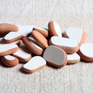 May include: A pile of small, smooth stones in terracotta and white. The stones have a natural, earthy appearance, with a terracotta base and white painted tops. They are arranged on a textured, neutral-toned surface.