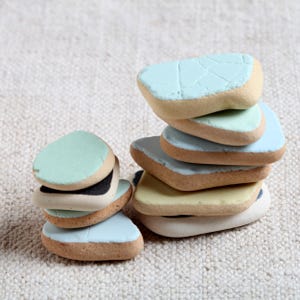 May include: A stack of painted ceramic stones in various pastel colors. The stones have a rounded, irregular shape with a light brown edge and a smooth, painted top in shades of blue, green, and yellow. The stones are stacked on a neutral-colored surface.