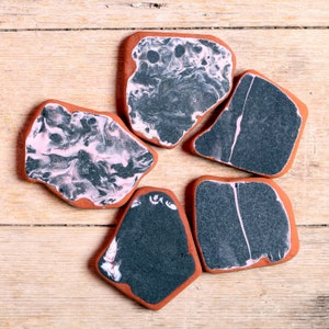 May include: Five irregularly shaped ceramic coasters with a reddish-brown border. The coasters feature a marbled design in shades of black, white, and pink. The coasters are arranged on a light-colored wooden surface.