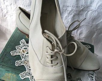 May include: A pair of off-white leather lace-up shoes with a rounded toe. The shoes are sitting on top of a stack of books with a lace doily underneath. The text "When Roses Bloom® WhenRosesBloomShop WRBshop" is visible in the image.