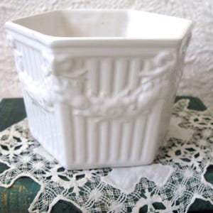 May include: A white ceramic planter with a hexagonal shape and vertical fluted sides. The exterior features a raised relief pattern of swags and garlands encircling the upper portion of the vessel, creating a textured and decorative appearance.