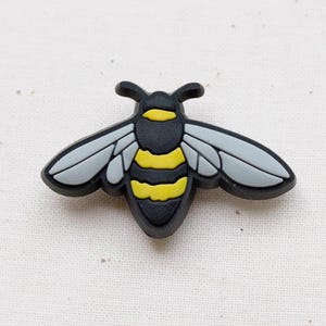 Puede incluir: Un dije con forma de abeja en negro, amarillo y gris. La abeja tiene alas grises, un abdomen a rayas amarillas y negras y una cabeza y tórax negros. El dije probablemente está hecho de goma o un material similar.