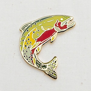 May include: A colorful enamel pin in the shape of a leaping trout. The fish features a yellow, green, red, and gold design with detailed scales and fins. The pin is likely for a jacket, backpack, or hat.