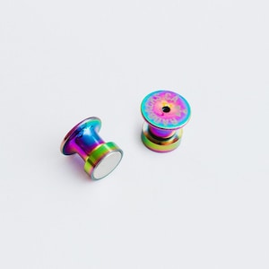 May include: Two iridescent magnetic stud earrings. The earrings have a rainbow finish with shades of purple, blue, green, and gold. The top of one earring has the text "LAS RAD". The earrings are on a white background.