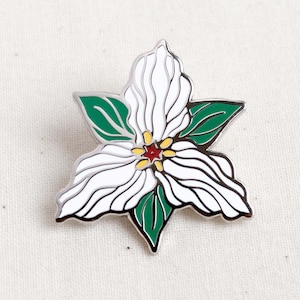 Op de afbeelding: Witte emaille pin met een groen en wit trillium bloeiende ontwerp. De bloem heeft een rood en geel centrum.