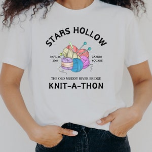 Könnte beinhalten: Weißes T-Shirt mit schwarzem Text, der "Stars Hollow" und "The Old Muddy River Bridge Knit-A-Thon" lautet. Der Text ist von einer Zeichnung von Wollknäueln und Stricknadeln umgeben.