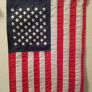 May include: A red, white, and blue American flag hanging on a white wall. The flag has 13 red and white stripes and a blue field with 50 white stars.