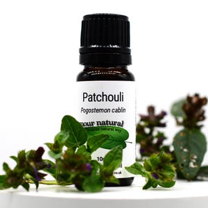 Puede incluir: Una pequeña botella de vidrio marrón de aceite esencial de Pachulí con una tapa negra. La etiqueta incluye el nombre, el nombre botánico y la frase "amour natural". Hojas verdes y flores moradas están dispuestas alrededor de la botella.