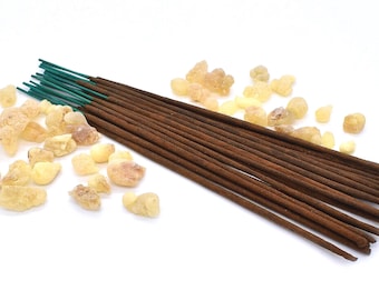 May include: A bundle of brown incense sticks with green tips, arranged on a white surface. Scattered around the sticks are small, pale yellow resin pieces.