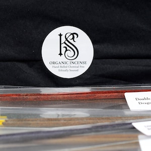May include: Incense sticks in a clear plastic package. The sticks are various colors, including red, yellow, and brown. A white label reads "Double Absolute Dragons Blood." A round sticker with the text "Organic Incense" is visible.
