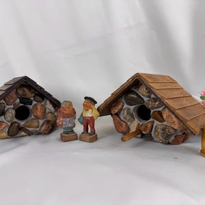 May include: Two stone birdhouses with brown shingled roofs, featuring round entry holes. Two small figurines of people stand between the birdhouses. A small wooden barrel holds pink flowers. The birdhouses are made of various coloured stones.