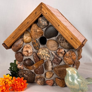 May include: A stone-clad birdhouse with a wooden roof. The birdhouse is made from a variety of brown, tan, and grey stones. A small ceramic bird and orange flowers are in the foreground, adding a decorative touch.
