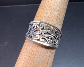 May include: A silver ring with an openwork design featuring swirling floral patterns. The ring is wide and sits on a wooden ring mandrel. The intricate details of the ring are highlighted against the neutral background.